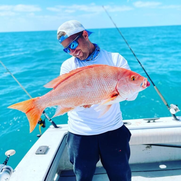 A man stands on a boat under a bright blue sky, holding a large red snapper fish with a big smile on his face. He is wearing a white long-sleeve shirt, a white cap, a blue neck gaiter, and sunglasses. The turquoise waters of the ocean are in the background, with fishing rods visible on the boat. This fish was caught on a fishing charter with Fish Daily Charters, specializing in "Half day fishing charters Florida Keys."