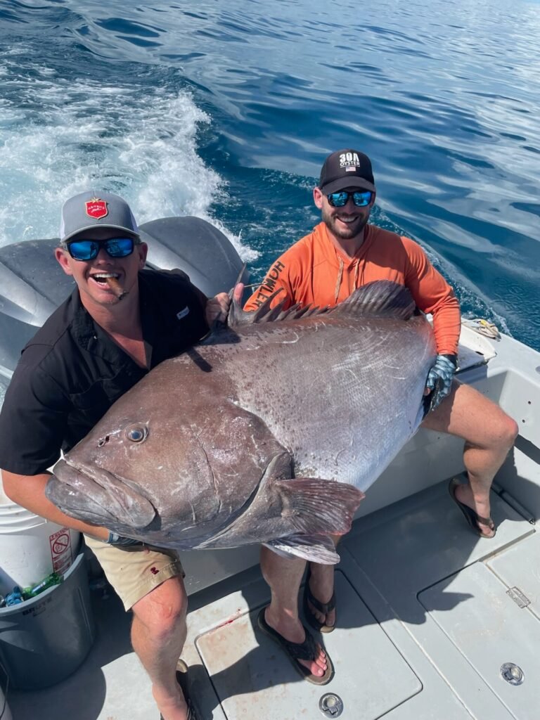 Two men sit on a boat holding an enormous grouper fish between them. The man on the left, wearing a black shirt and a cap, has a cigar in his mouth and a big smile. The man on the right is wearing an orange shirt, a black cap, and sunglasses, also smiling widely. The ocean is visible behind them, with the boat in motion. This impressive catch was made on a fishing charter with Fish Daily Charters, specializing in "Grouper fishing charters in Key West."