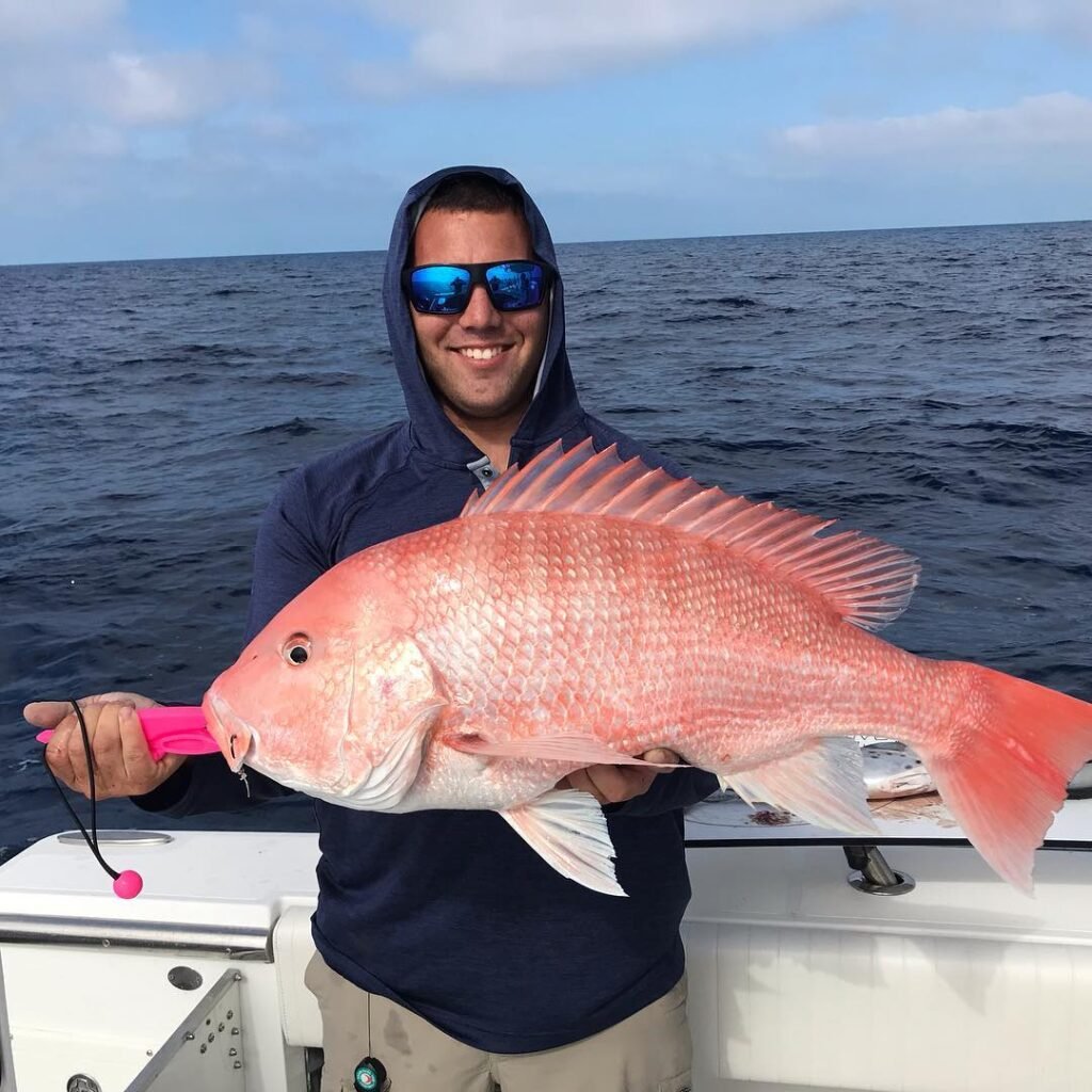 A man stands on a boat holding a large red snapper fish with a bright pink lip grip. He is wearing a blue hoodie, beige pants, and sunglasses, smiling broadly. The ocean and a partly cloudy sky are visible in the background. This snapper was caught on a fishing charter with Fish Daily Charters, specializing in "Key West Snapper Fishing."