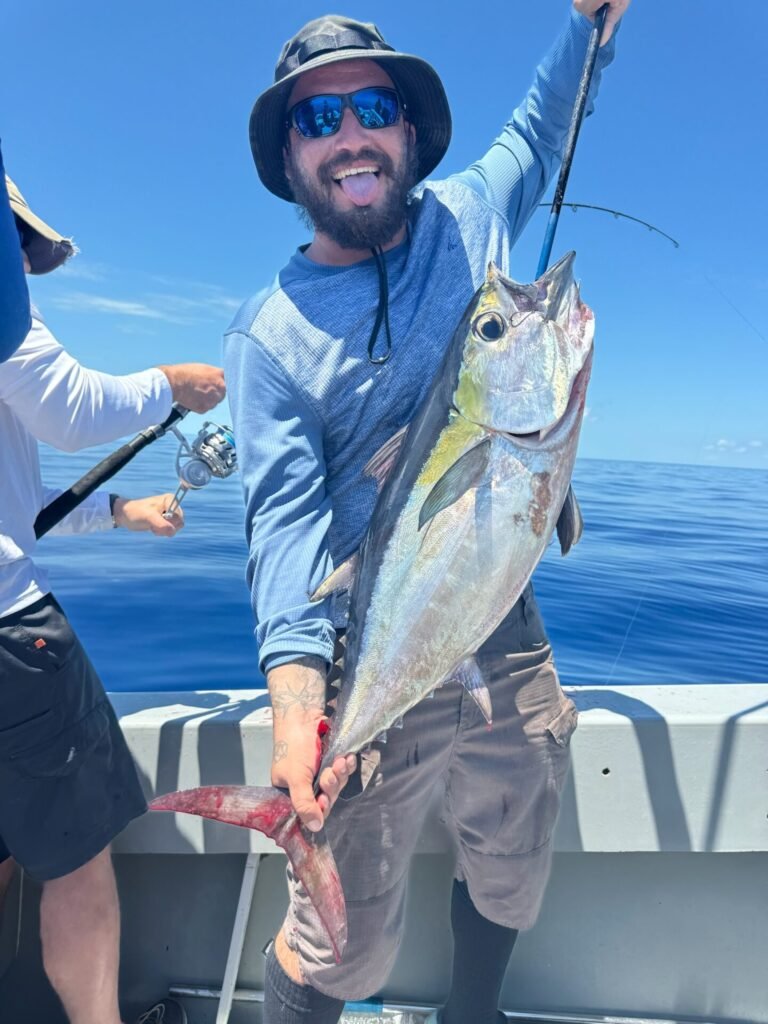 A man stands on a boat under a clear blue sky, proudly holding up a freshly caught tuna by the tail. He is wearing a light blue long-sleeve shirt, grey shorts, black sunglasses, and a wide-brimmed hat, with a big smile and his tongue playfully sticking out. Another person in the background is also fishing. This tuna was caught on a fishing charter with Fish Daily Charters, specializing in "Key West Tuna Fishing."