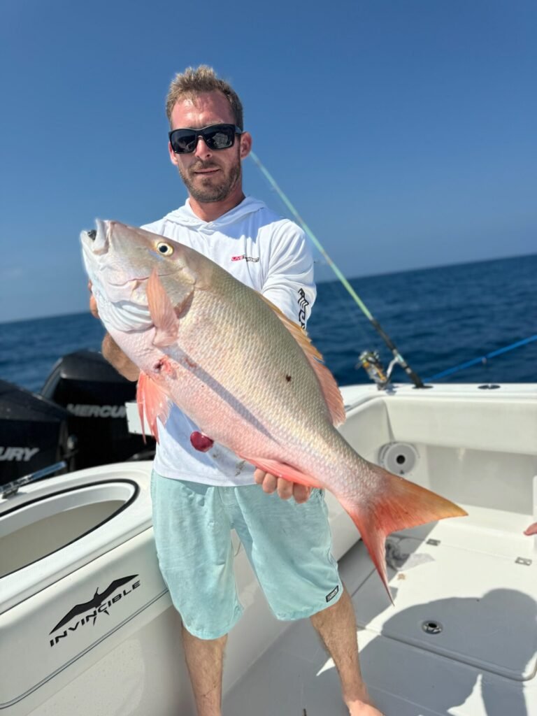 A man stands on a boat under a clear blue sky, holding a large red snapper fish. He is wearing a white long-sleeve hoodie, light blue shorts, and dark sunglasses. The ocean is visible in the background, along with fishing rods. This snapper was caught on a fishing charter with Fish Daily Charters, specializing in "Snapper Fishing Charters in Key West."