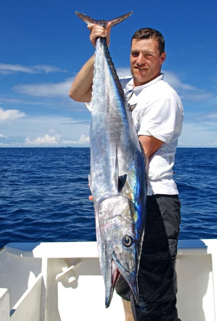 Angler proudly holding a large, gleaming wahoo on a boat during a Key West Wahoo Fishing Charter with Fish Daily Charters, with the clear blue sky and ocean as a backdrop.