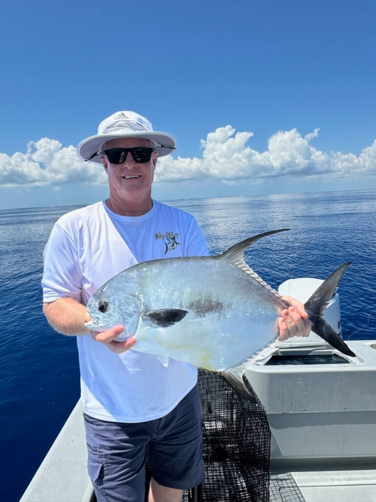 Smiling angler proudly displaying a large permit fish on a boat during a Permit Fishing Charter in Key West with Fish Daily Charters, under a bright blue sky with fluffy clouds.