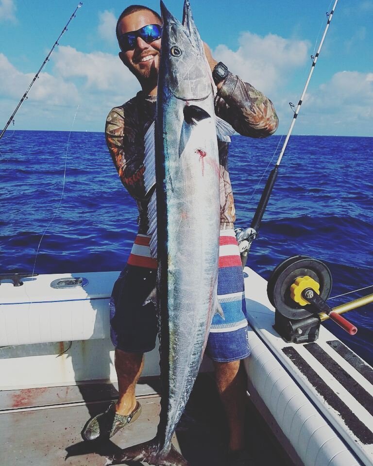 Angler smiling while holding a large wahoo on a boat during a Wahoo Fishing Charter in Key West with Fish Daily Charters, with the deep blue ocean and clear sky in the background.