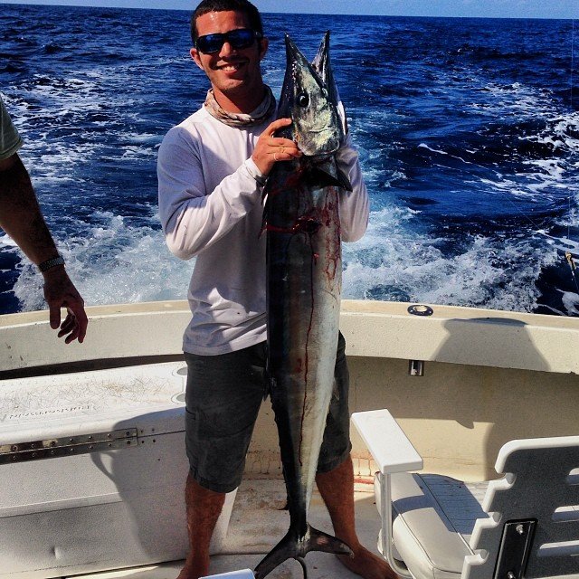 Angler proudly holding a large wahoo on a boat during a Wahoo Fishing Charter in Key West with Fish Daily Charters, with the wake of the boat and the deep blue ocean in the background.
