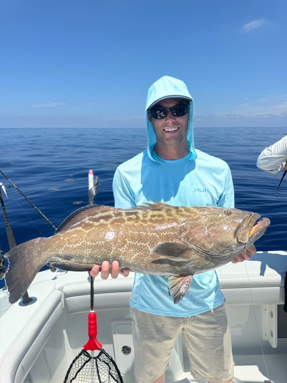 Grouper Fishing charter Key West A man stands on a boat under a clear blue sky, holding a large, patterned grouper fish. He is wearing a light blue long-sleeve shirt with a hood, sunglasses, and beige shorts. The calm ocean serves as a backdrop. This grouper was caught on a fishing charter with Fish Daily Charters, specializing in "Grouper fishing charter Key West."