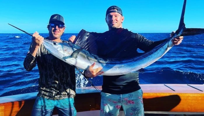 Key West Sailfish Fishing Two anglers proudly holding a sailfish on a boat during a successful Key West Sailfish Fishing trip with Fish Daily Charters, with the deep blue ocean and clear sky in the background.