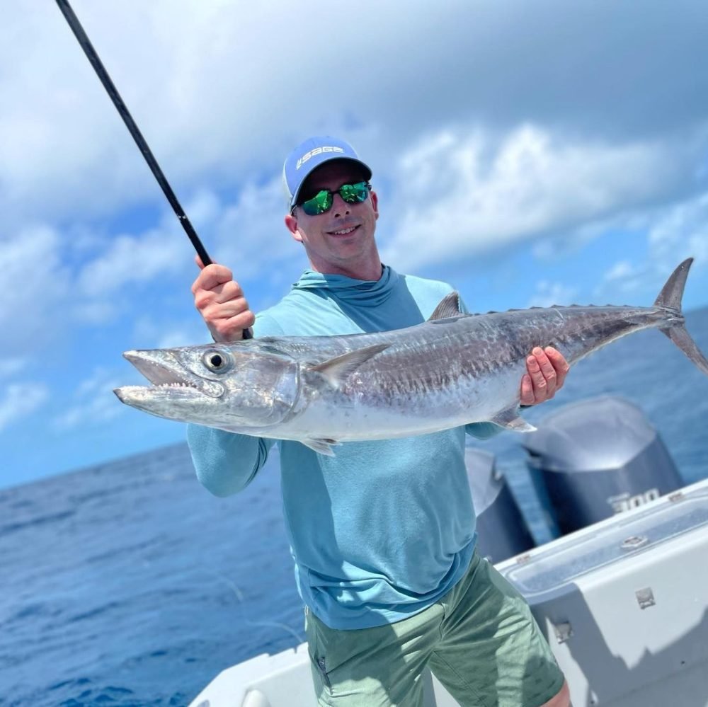 King Mackerel Fishing Charters in Key West Angler smiling while holding a large king mackerel on a boat during a King Mackerel Fishing Charter in Key West with Fish Daily Charters, with the ocean and a partly cloudy sky in the background.