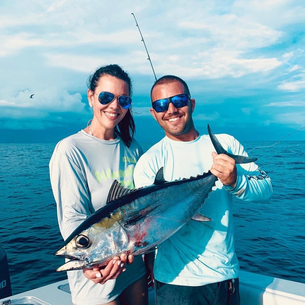 Tuna Fishing in key west A man and woman stand on a boat against a backdrop of a cloudy sky and calm ocean waters, both wearing light-colored long-sleeve shirts and sunglasses. They are smiling and holding a freshly caught tuna fish together, each gripping one end of the fish. This tuna was caught on a fishing charter with Fish Daily Charters, specializing in "Tuna Fishing in Key West."